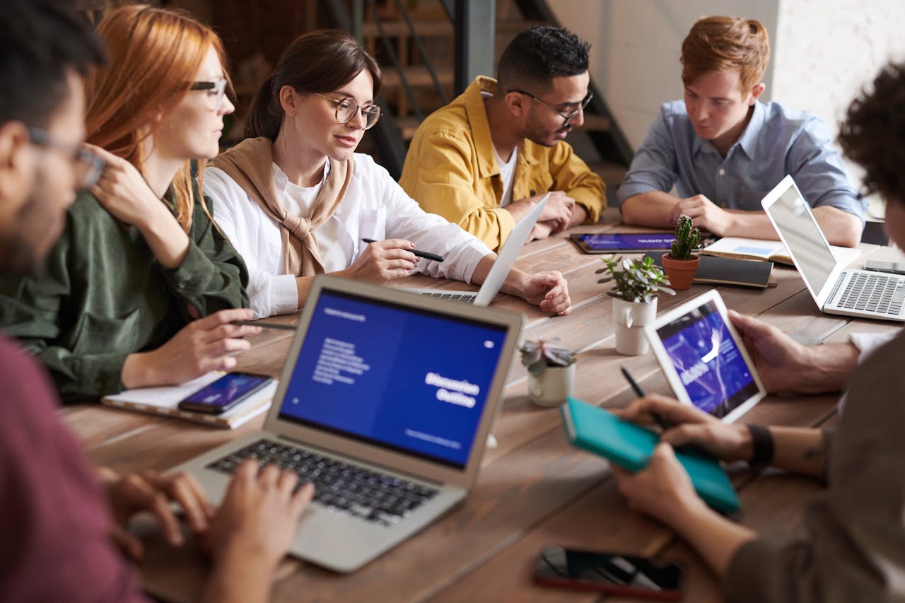 Group of people working together in an office meeting, collaborating with laptops, tablets, and notebooks.